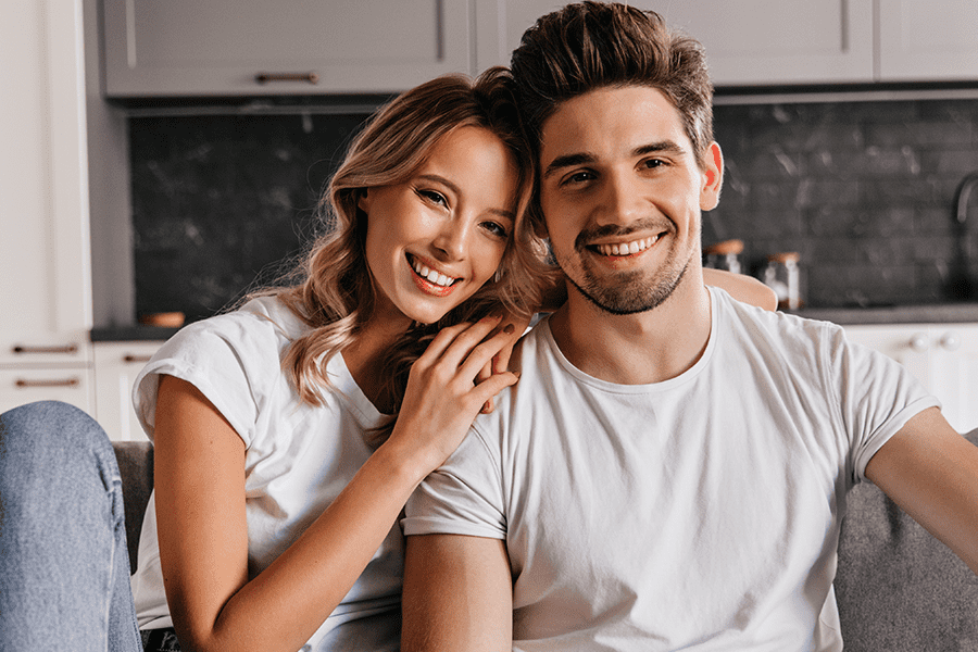 Young couple poses on a grey couch with kitchen cabinets behind them.
